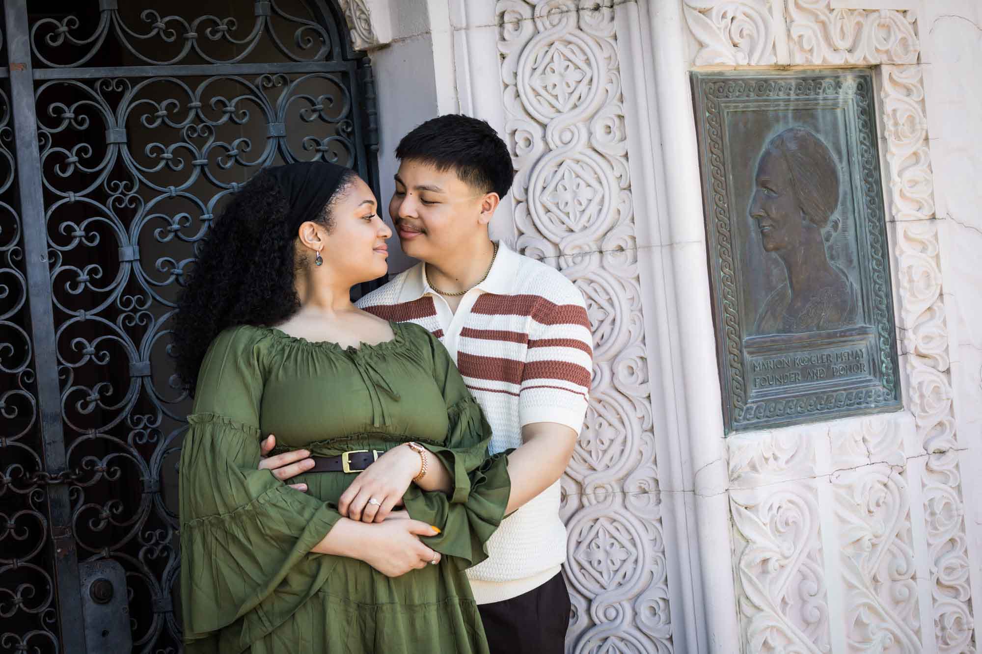 Man hugging woman wearing green dress in front of intricately carved stone arch doorway with wrought iron door and copper plaque for an article on Venice Canals engagement photos