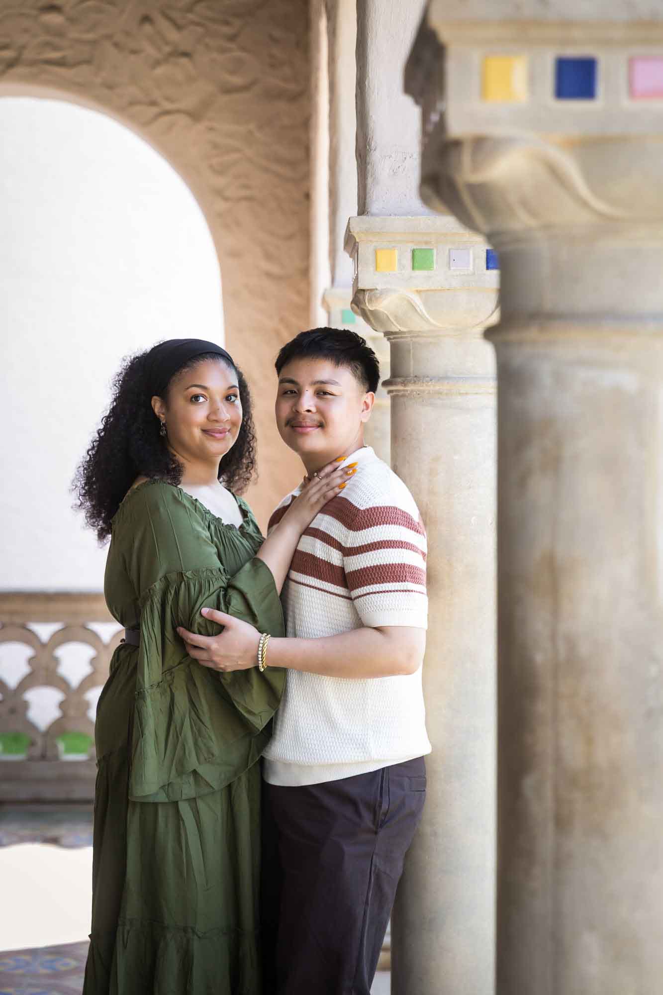 Woman wearing green dress hugging man resting against column with colorful mosaics at top for an article on Venice Canals engagement photos