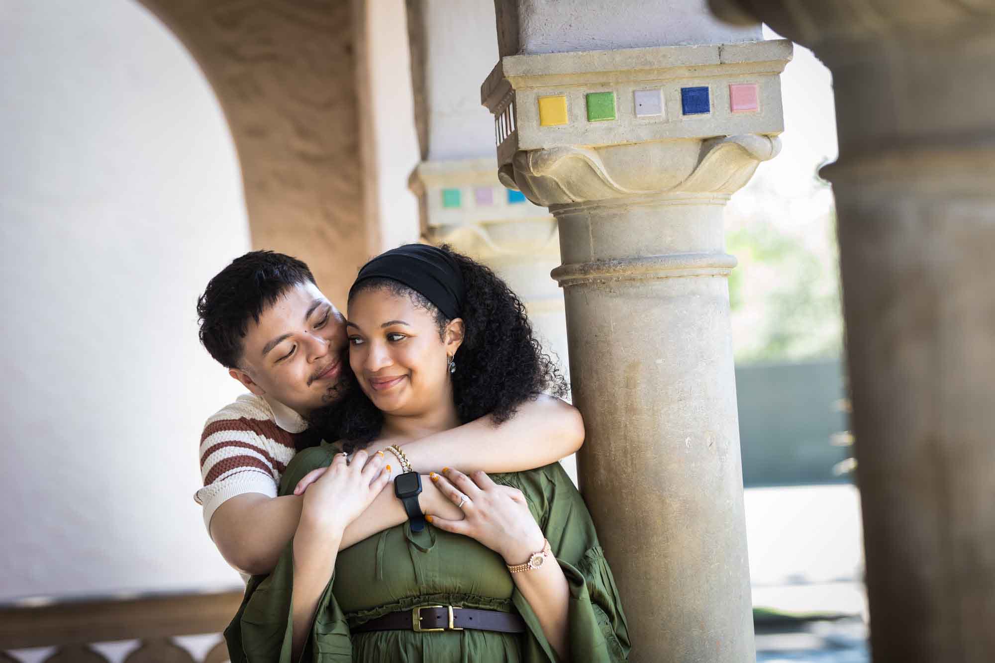 Man hugging woman wearing green dress resting against column with colorful mosaics at top for an article on Venice Canals engagement photos