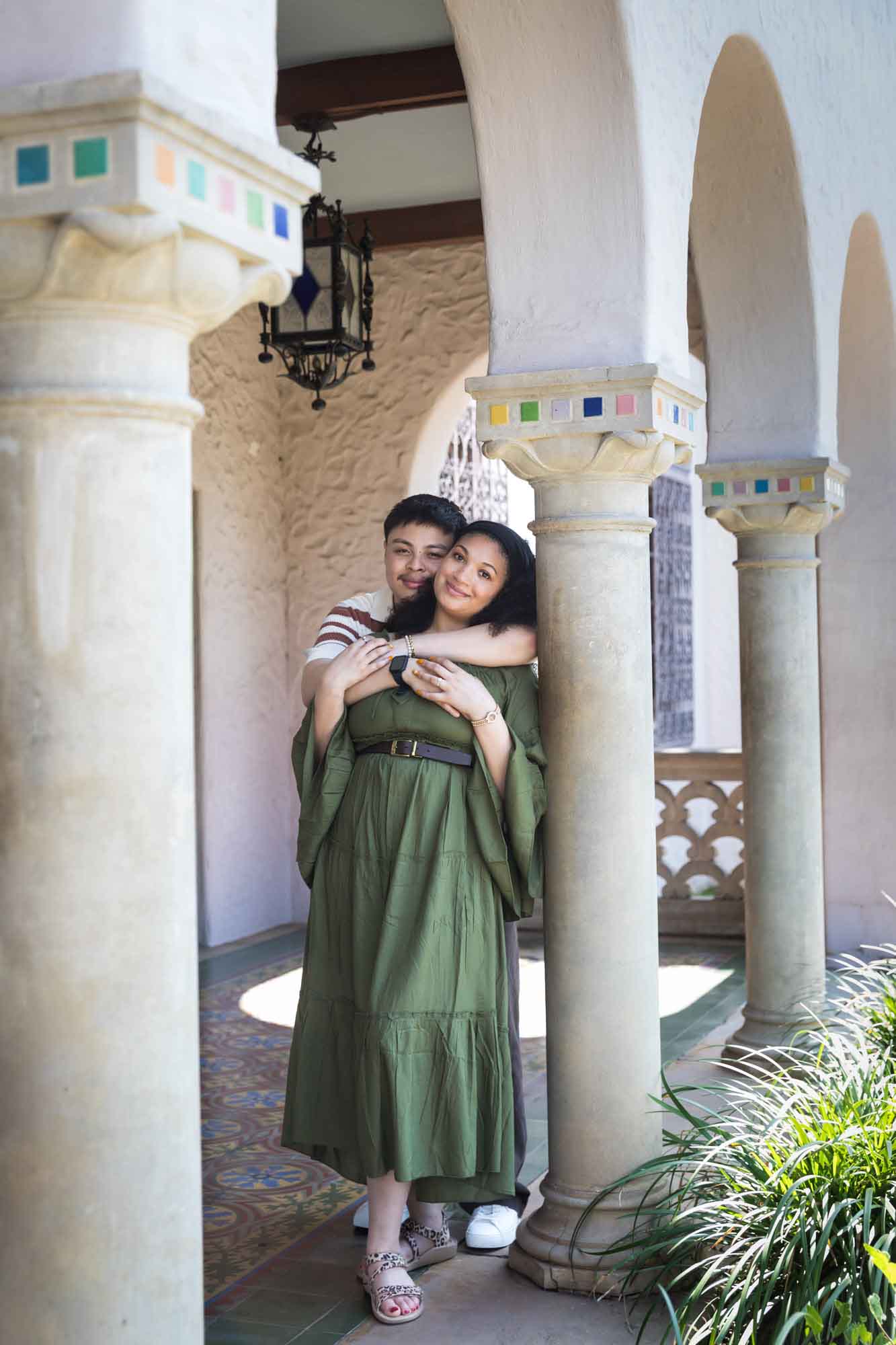 Man hugging woman wearing green dress resting against column with colorful mosaics at top for an article on Venice Canals engagement photos