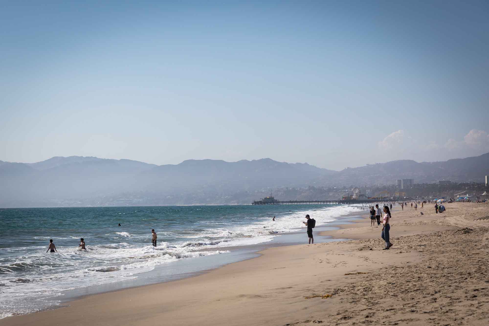 People walking into the ocean and along the sand in Venice Beach with buildings and mountains in the background for an article on Venice Canals engagement photos