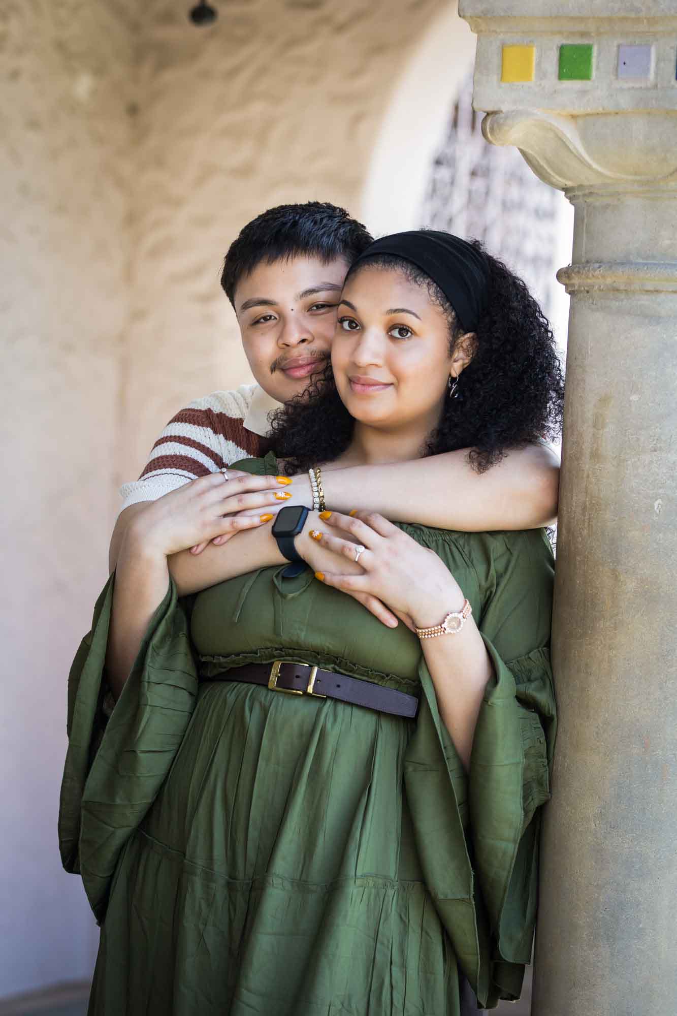 Man hugging woman wearing green dress resting against column with colorful mosaics at top for an article on Venice Canals engagement photos