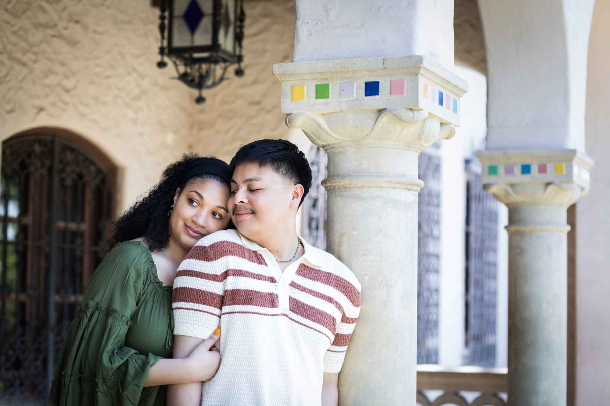 Woman wearing green dress hugging arm of man resting against column with colorful mosaics at top for an article on Venice Canals engagement photos