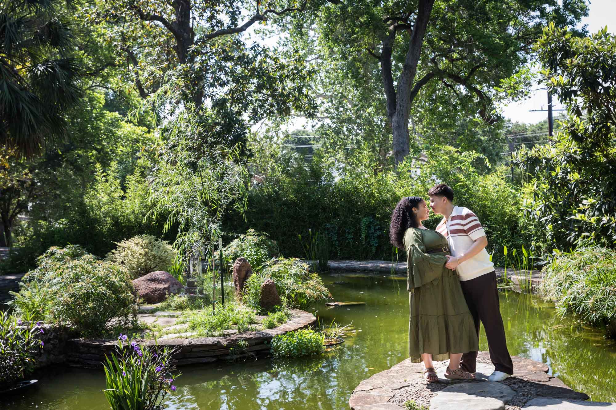 Woman wearing green dress standing at angle with man on stone island in garden lake for an article on Venice Canals engagement photos