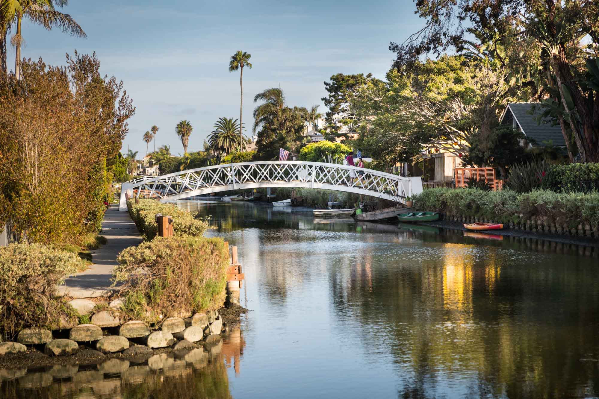 White footbridge over a canal with boats, trees, and small docks on the side along the Venice Canals in Los Angeles for an article on Venice Canals engagement photos