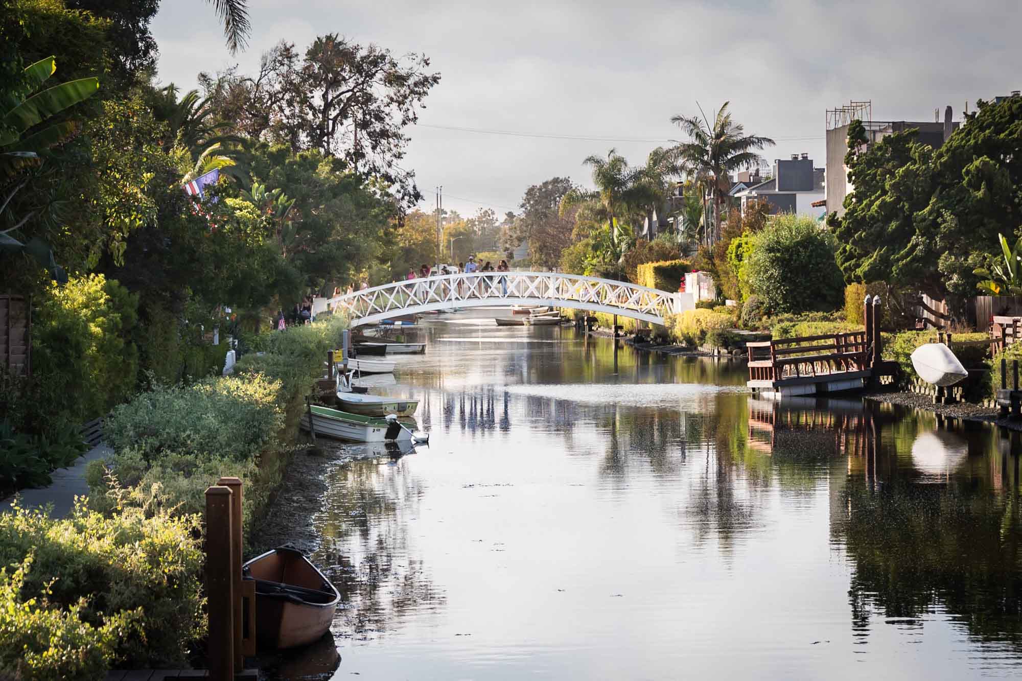 White footbridge over a canal with boats, trees, and small docks on the side along the Venice Canals in Los Angeles for an article on Venice Canals engagement photos