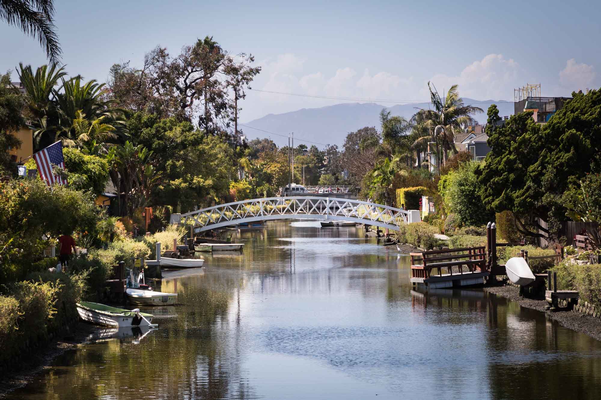 White footbridge over a canal with boats, trees, and small docks on the side along the Venice Canals in Los Angeles for an article on Venice Canals engagement photos