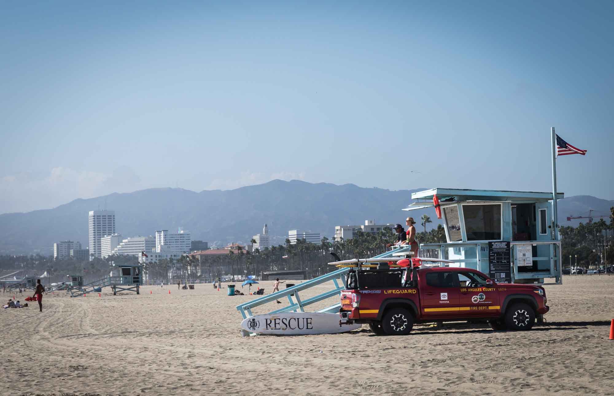 A male and female lifeguard watching from an aqua colored lifeguard station behind a red lifeguard truck parked on the sand in Venice Beach for an article on Venice Canals engagement photos