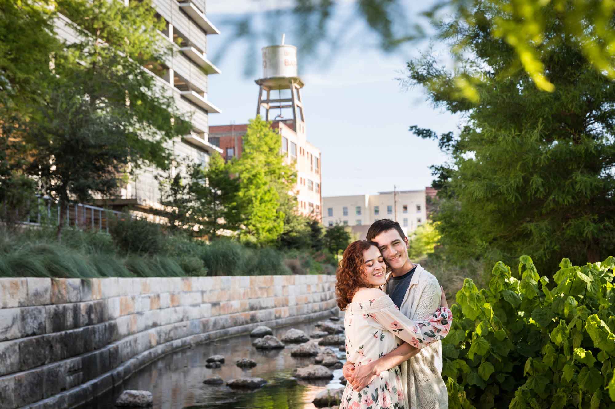 Couple hugging in front of creek and brick wall with water container on top of building and parking deck in background for an article on Walt Disney Concert Hall photos