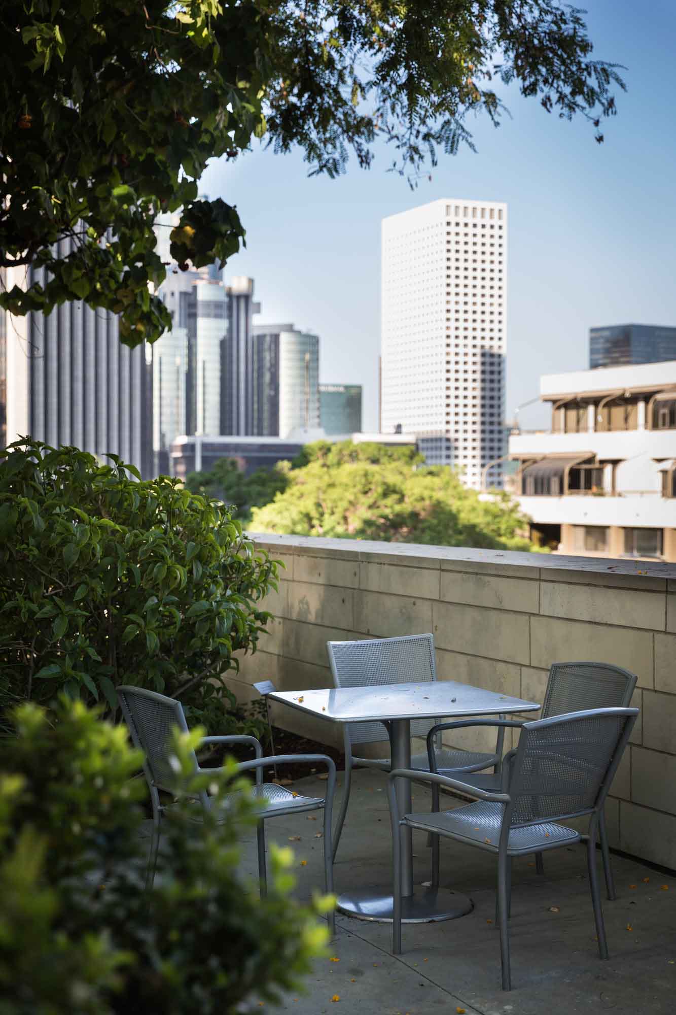 Silver table and four chairs next to two green bushes and stone wall with skyscrapers of downtown Los Angeles in the background for an article on Walt Disney Concert Hall photos