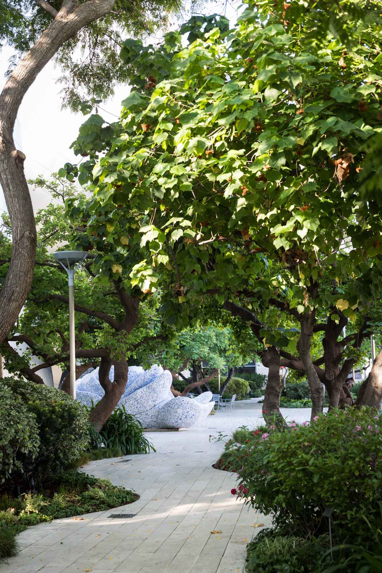 Undulating white brick pathway leading through trees and bushes in Blue Ribbon Garden leading to mosaic fountain for an article on Walt Disney Concert Hall photos