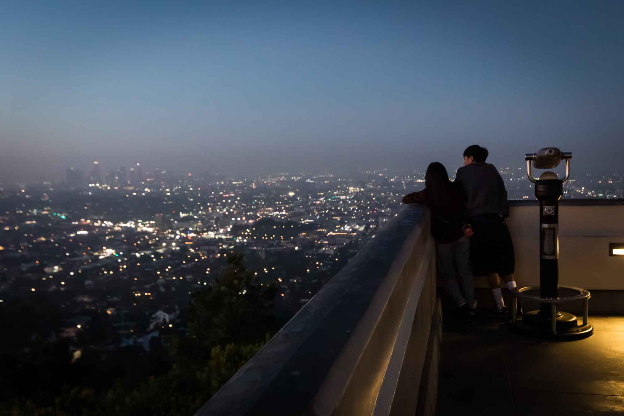 Couple standing on East Terrace beside binoculars looking at Los Angeles skyline before sunrise for an article on where to propose at the Griffith Observatory