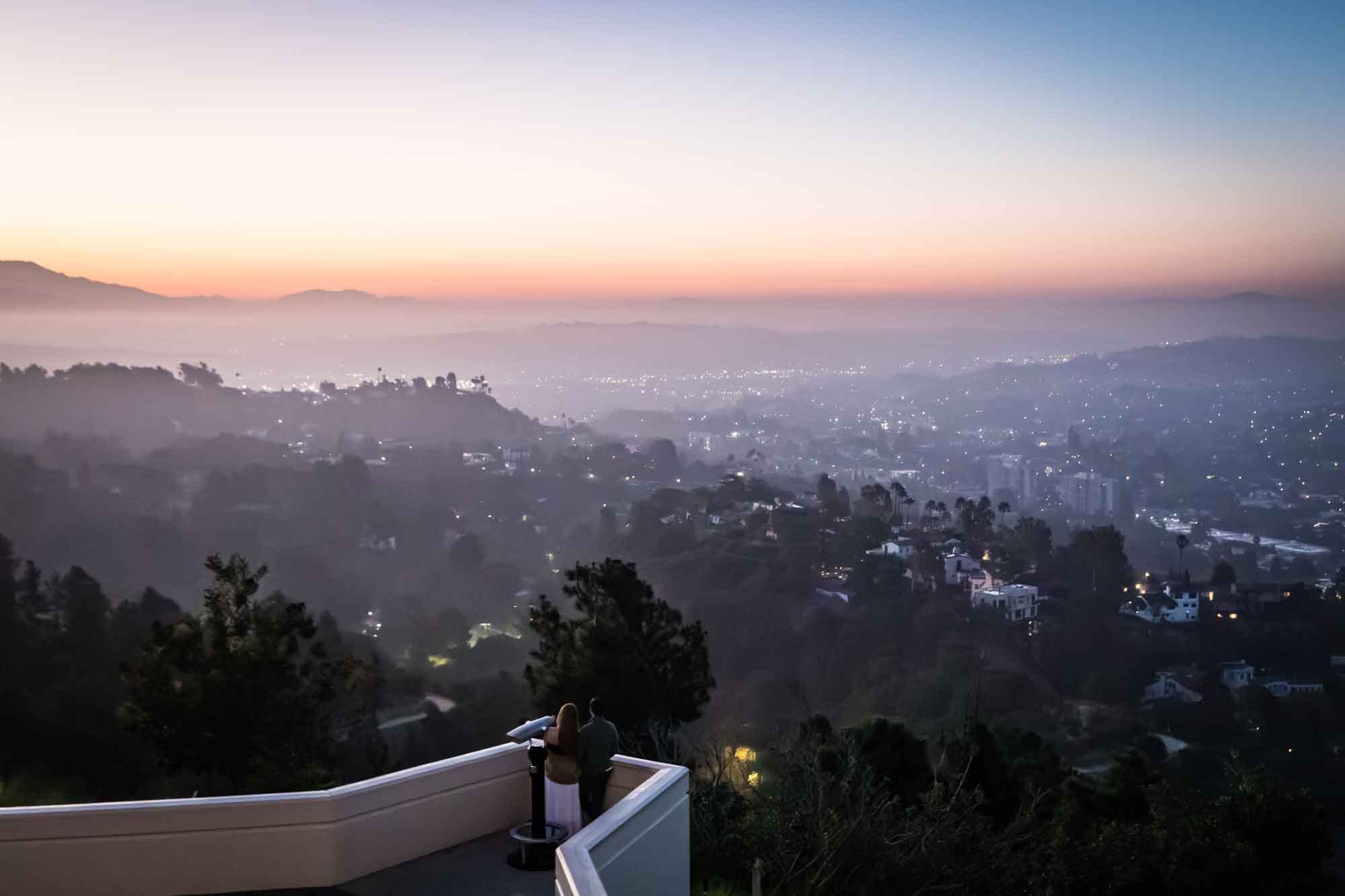 Couple standing on East Terrace by binoculars with Los Angeles skyline in background before sunrise for an article on where to propose at the Griffith Observatory