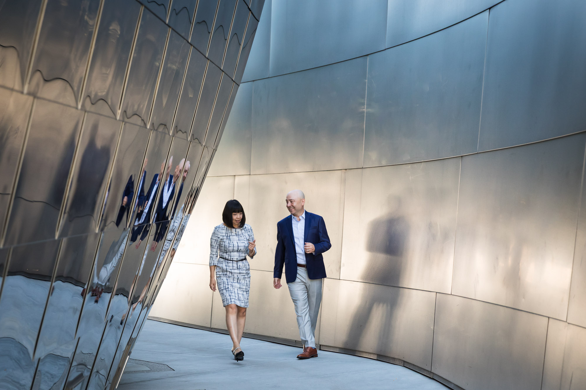 Businessman wearing blue jacket and khaki pants walking and talking with Asian American female colleague wearing dress in front of metal walls of Walt Disney Concert Hall for an article on how to prepare for a branding photo shoot