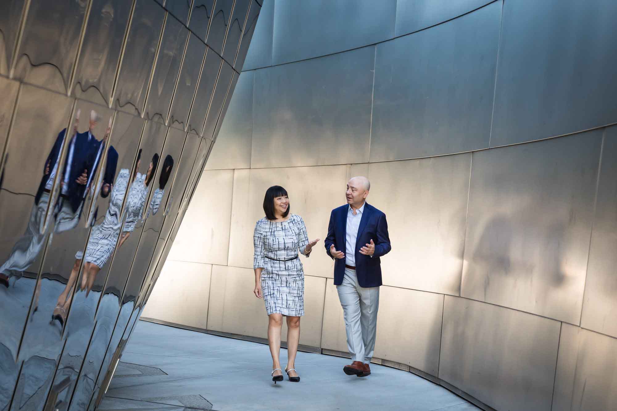 Businessman wearing blue jacket and khaki pants walking and talking with Asian American female colleague wearing dress in front of metal walls of Walt Disney Concert Hall for an article on how to prepare for a branding photo shoot