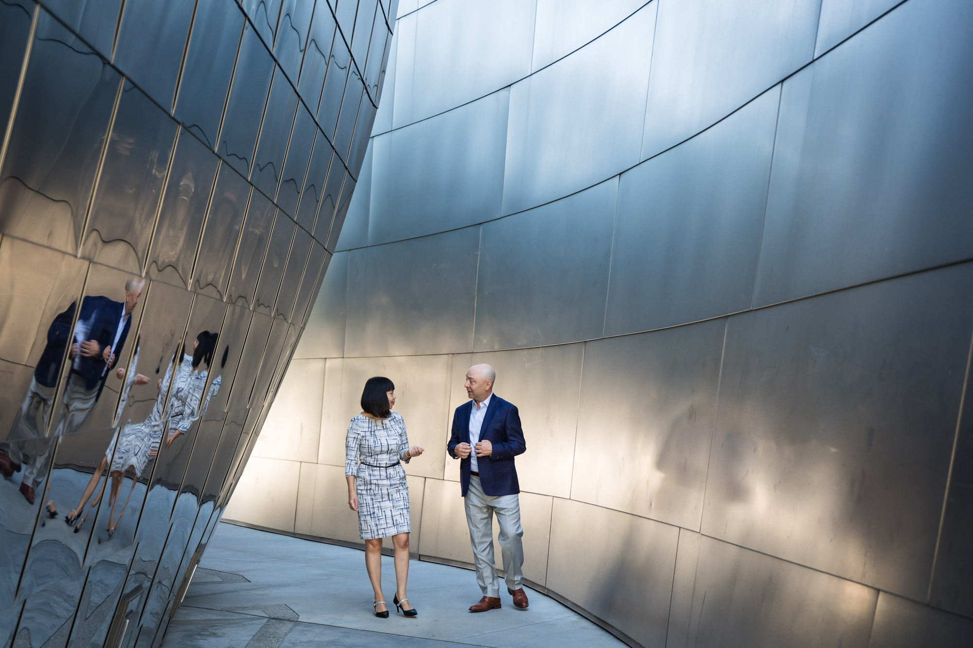 Businessman wearing blue jacket and khaki pants walking and talking with Asian American female colleague wearing dress in front of metal walls of Walt Disney Concert Hall for an article on how to prepare for a branding photo shoot