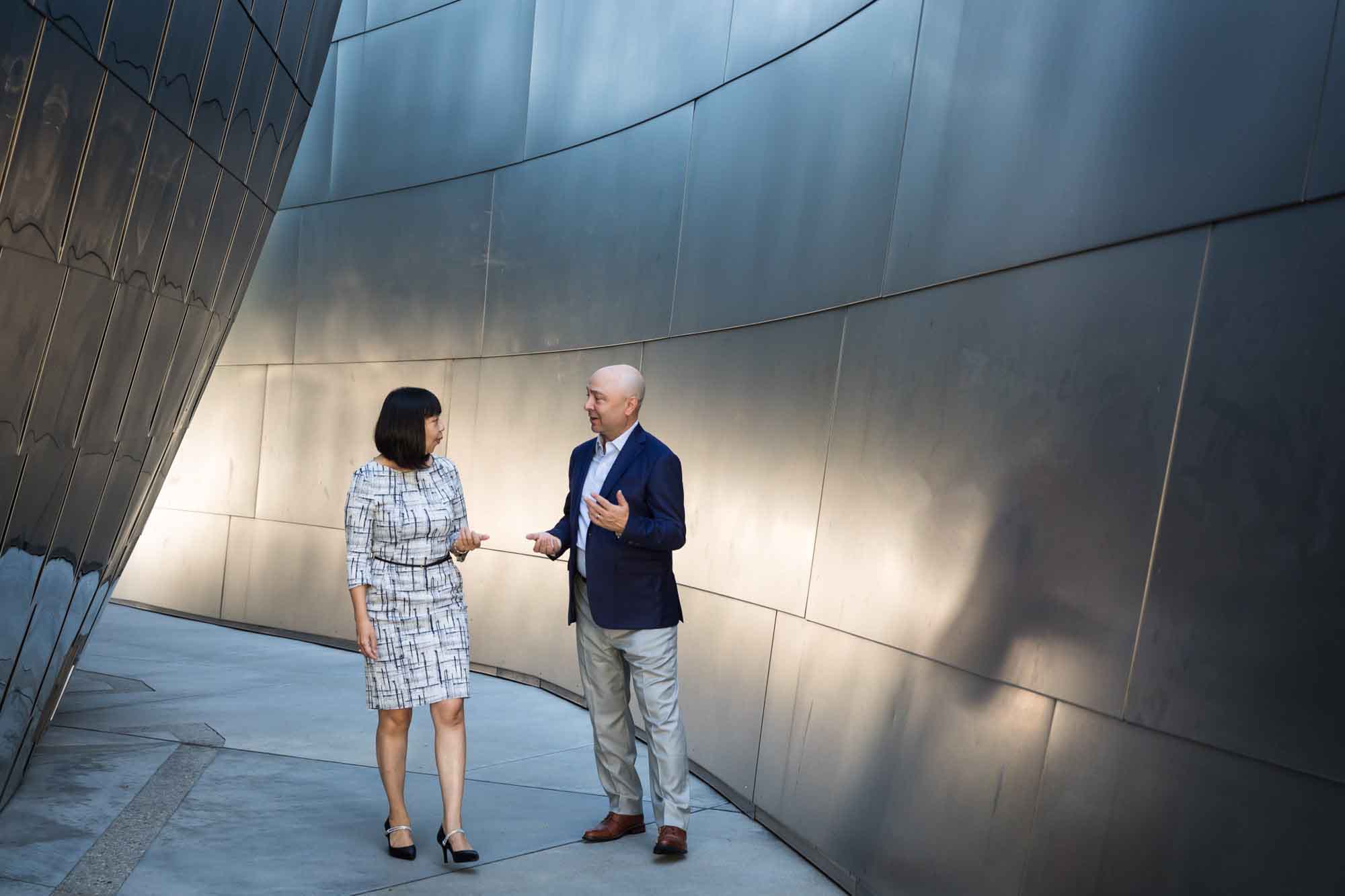 Businessman wearing blue jacket and khaki pants walking and talking with Asian American female colleague wearing dress in front of metal walls of Walt Disney Concert Hall for an article on how to prepare for a branding photo shoot