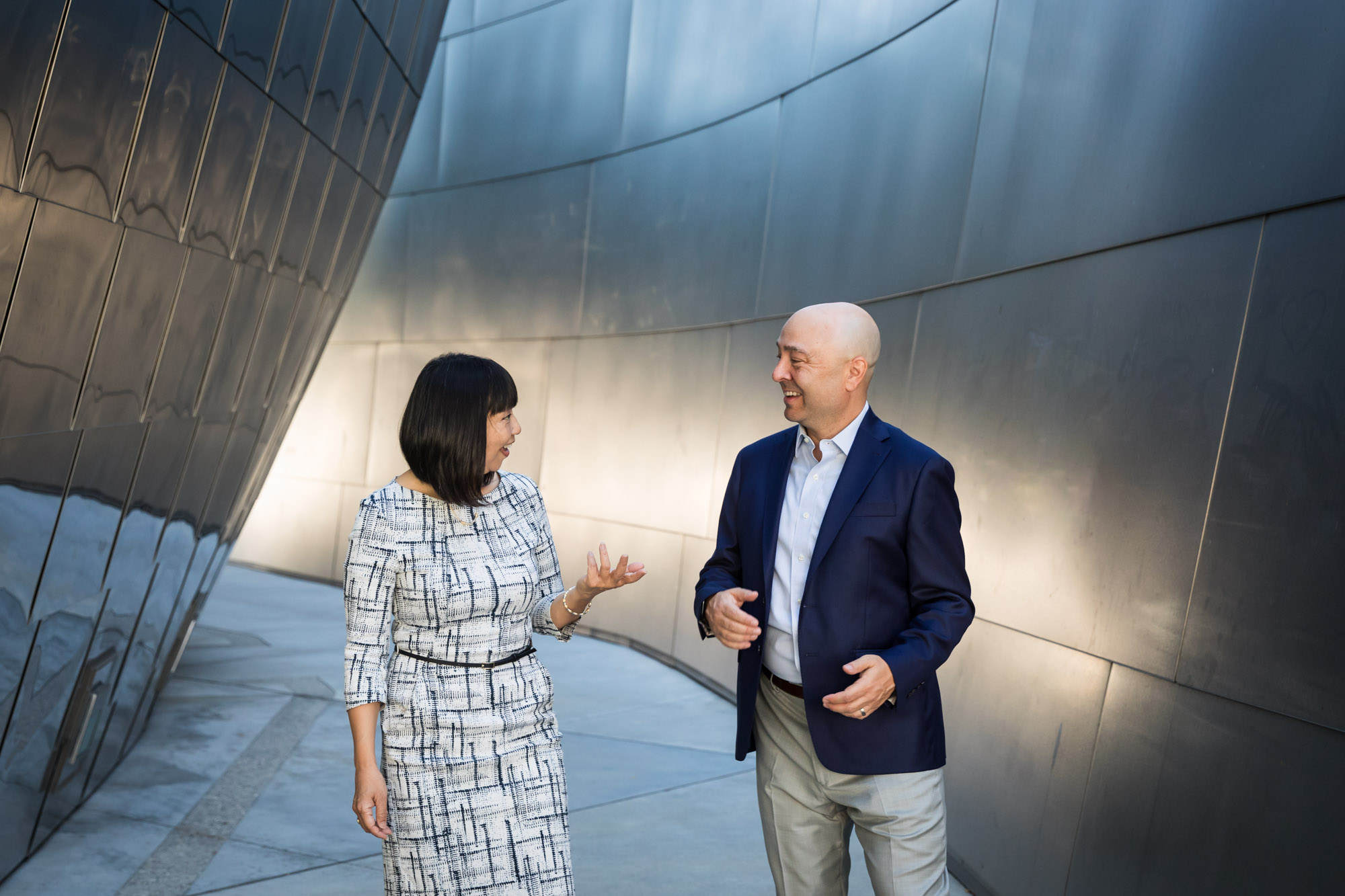 Businessman wearing blue jacket and khaki pants walking and talking with Asian American female colleague wearing dress in front of metal walls of Walt Disney Concert Hall for an article on how to prepare for a branding photo shoot