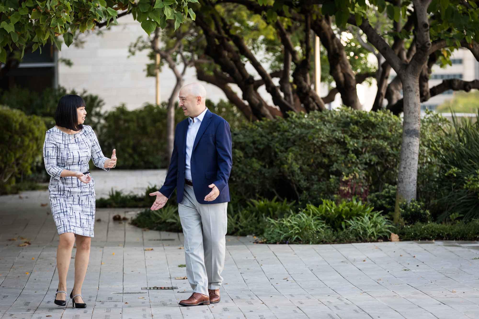 Businessman wearing blue jacket and khaki pants walking and talking with Asian American female colleague wearing dress in Blue Ribbon Garden for an article on how to prepare for a branding photo shoot