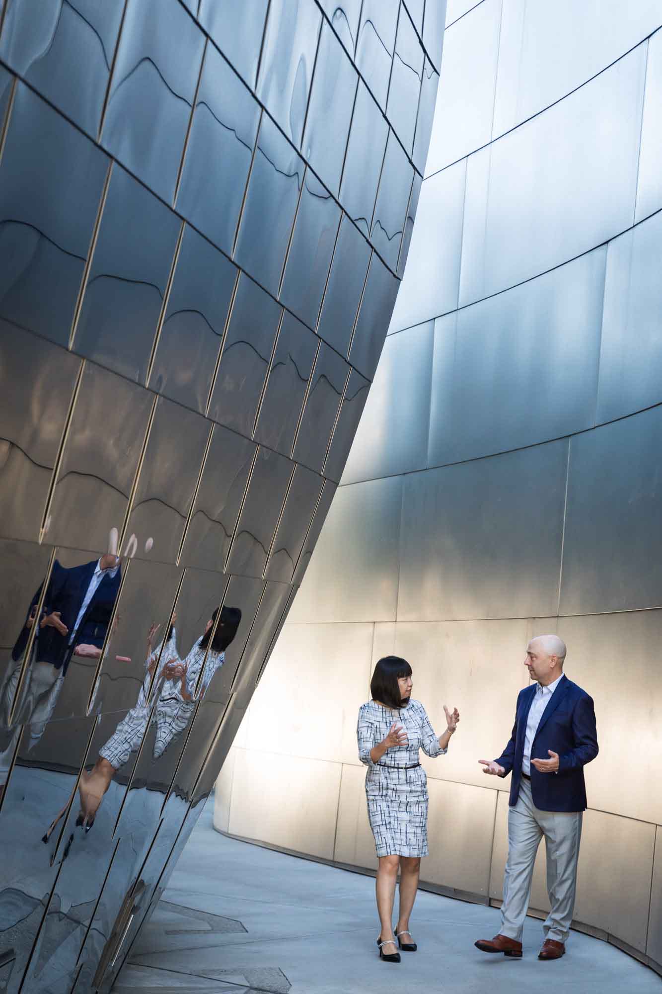 Businessman wearing blue jacket and khaki pants walking and talking with Asian American female colleague wearing dress in front of metal walls of Walt Disney Concert Hall for an article on how to prepare for a branding photo shoot