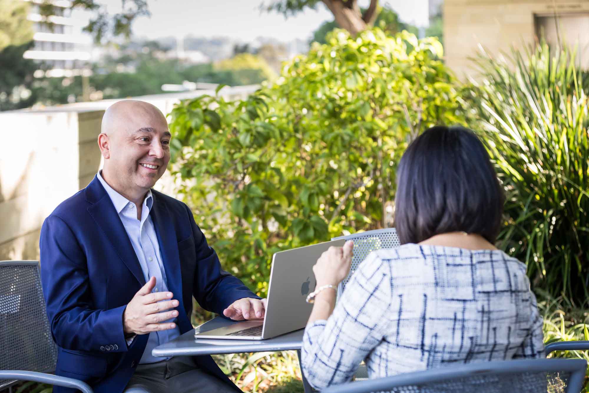 Businessman wearing blue jacket and khaki pants seated and talking with Asian American female colleague wearing dress in Blue Ribbon Garden for an article on how to prepare for a branding photo shoot
