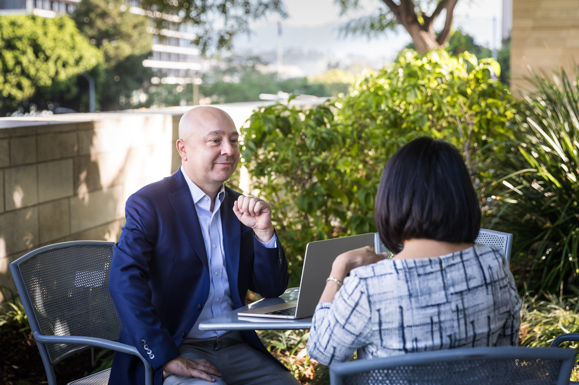 Businessman wearing blue jacket and khaki pants seated and talking with Asian American female colleague wearing dress in Blue Ribbon Garden for an article on how to prepare for a branding photo shoot