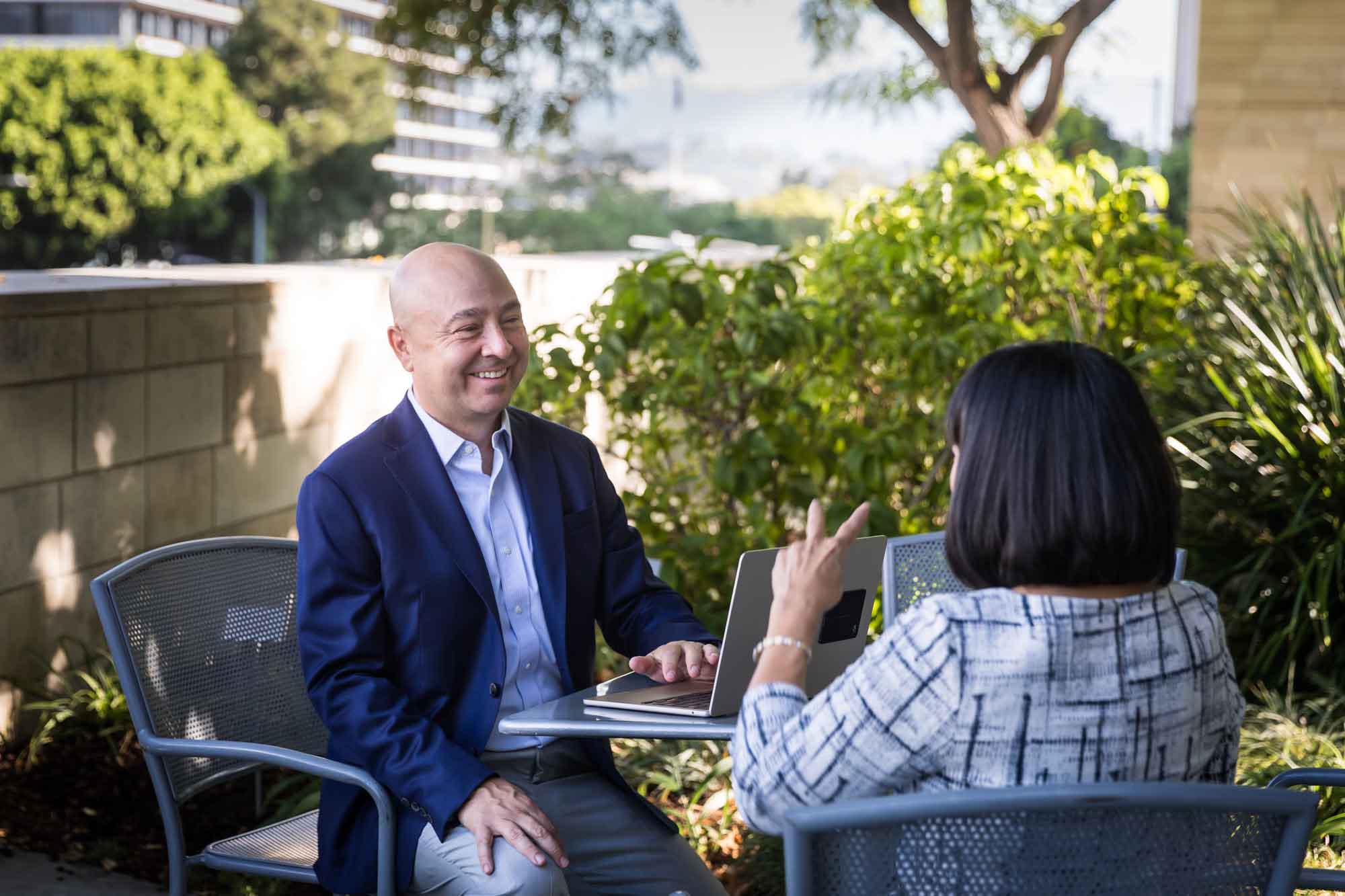 Businessman wearing blue jacket and khaki pants seated and talking with Asian American female colleague wearing dress in Blue Ribbon Garden for an article on how to prepare for a branding photo shoot