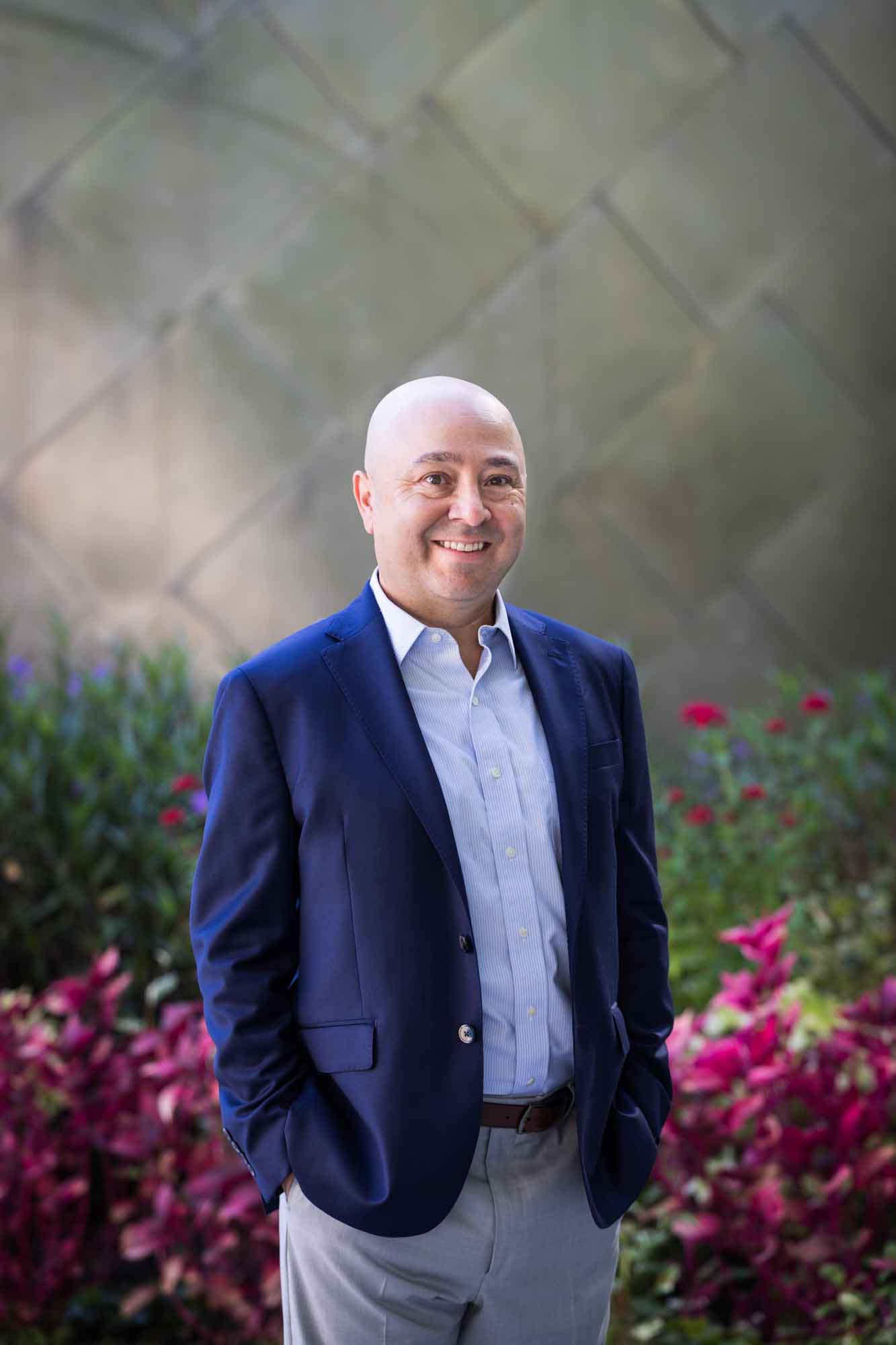 Executive portrait of businessman wearing blue jacket and khaki pants standing with hands in pockets in front of maroon flowers and metal wall for an article on how to prepare for a branding photo shoot