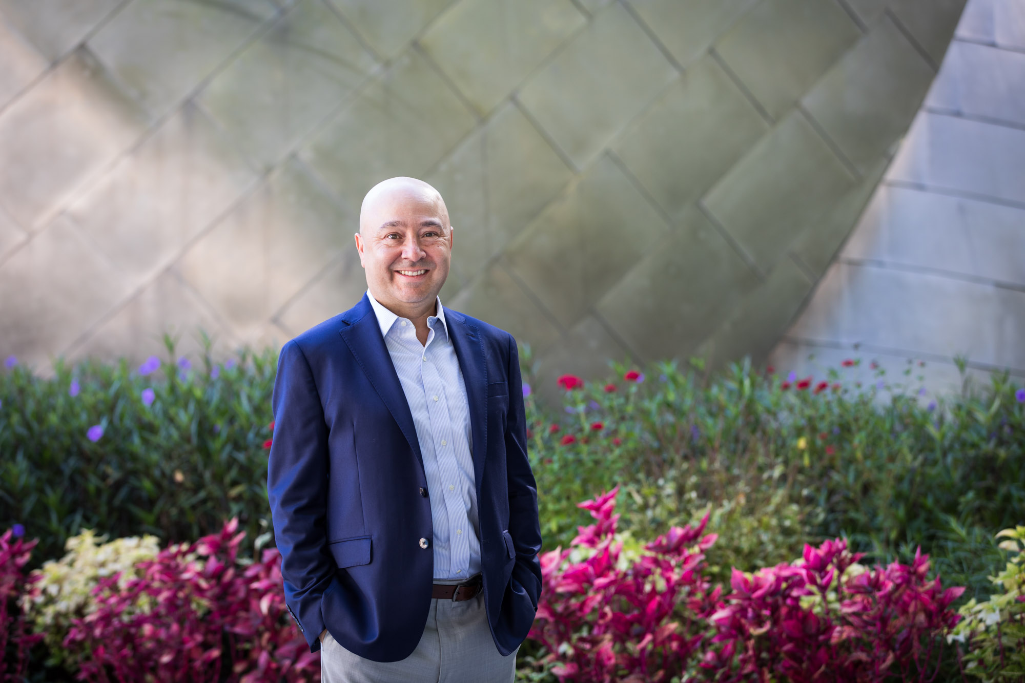 Executive portrait of businessman wearing blue jacket and khaki pants standing with hands in pockets in front of maroon flowers and metal wall for an article on how to prepare for a branding photo shoot