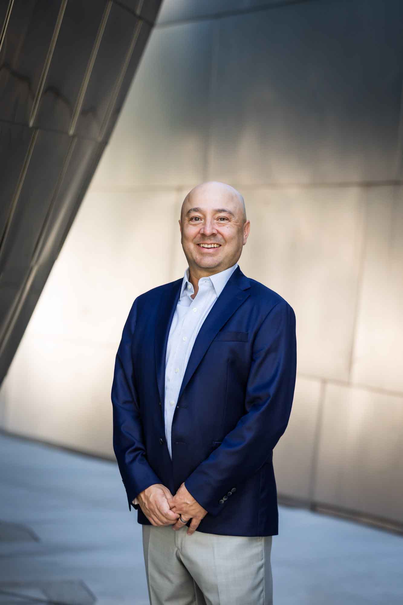 Executive portrait of businessman wearing blue jacket and khaki pants standing with hands in pockets in front of metal wall for an article on how to prepare for a branding photo shoot
