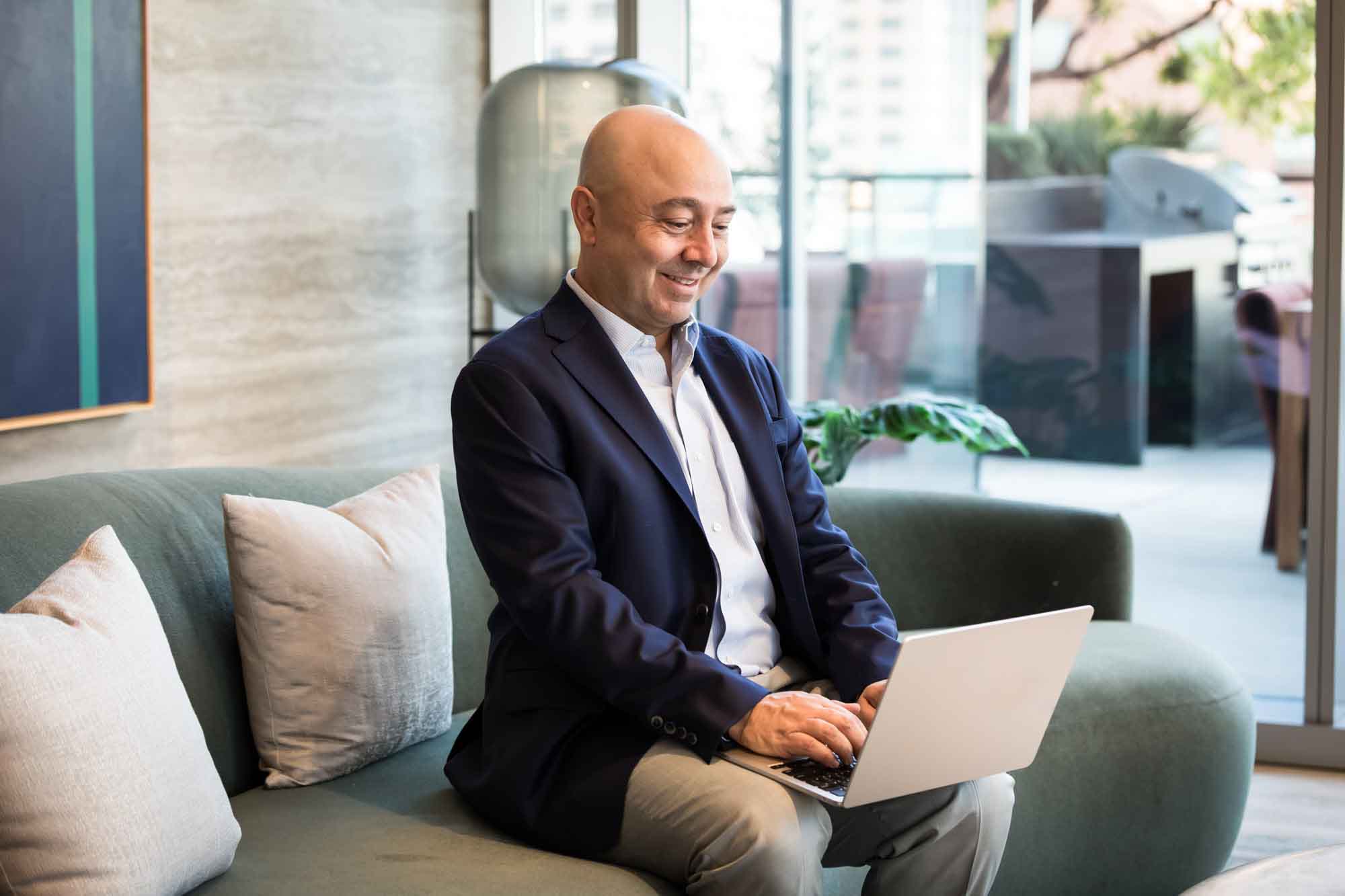 Executive portrait of businessman wearing blue blazer and blue and white shirt seated on grey couch typing on laptop in front of window for an article on how to prepare for a branding photo shoot