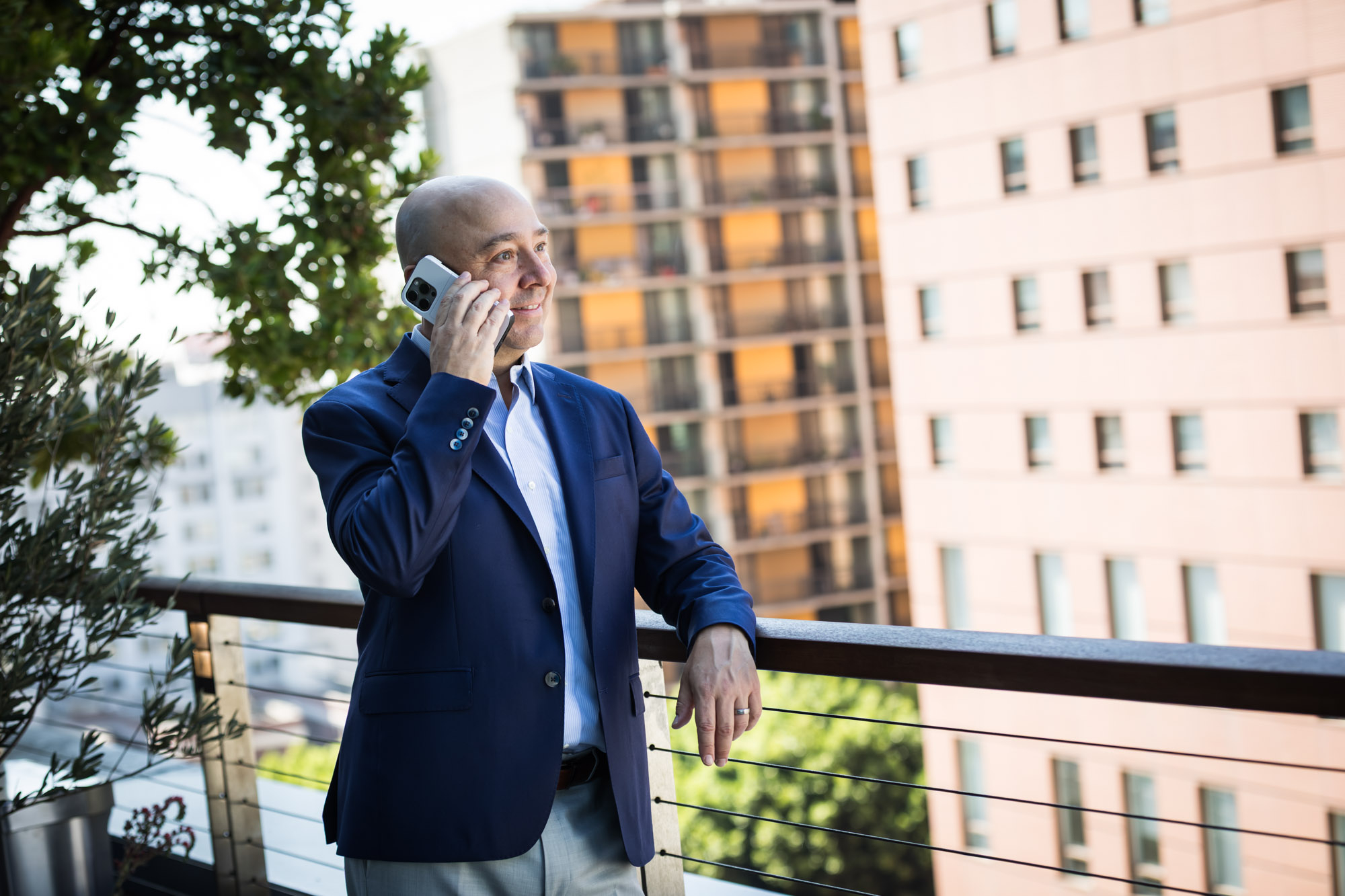 Executive portrait of businessman wearing blue jacket and khaki pants talking on phone and leaning against railing with apartment building in background for an article on how to prepare for a branding photo shoot