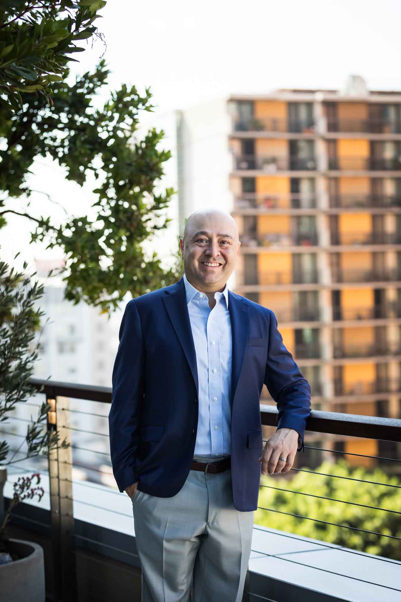 Executive portrait of businessman wearing blue jacket and khaki pants with hand in pocket and leaning against railing with apartment building in background for an article on how to prepare for a branding photo shoot