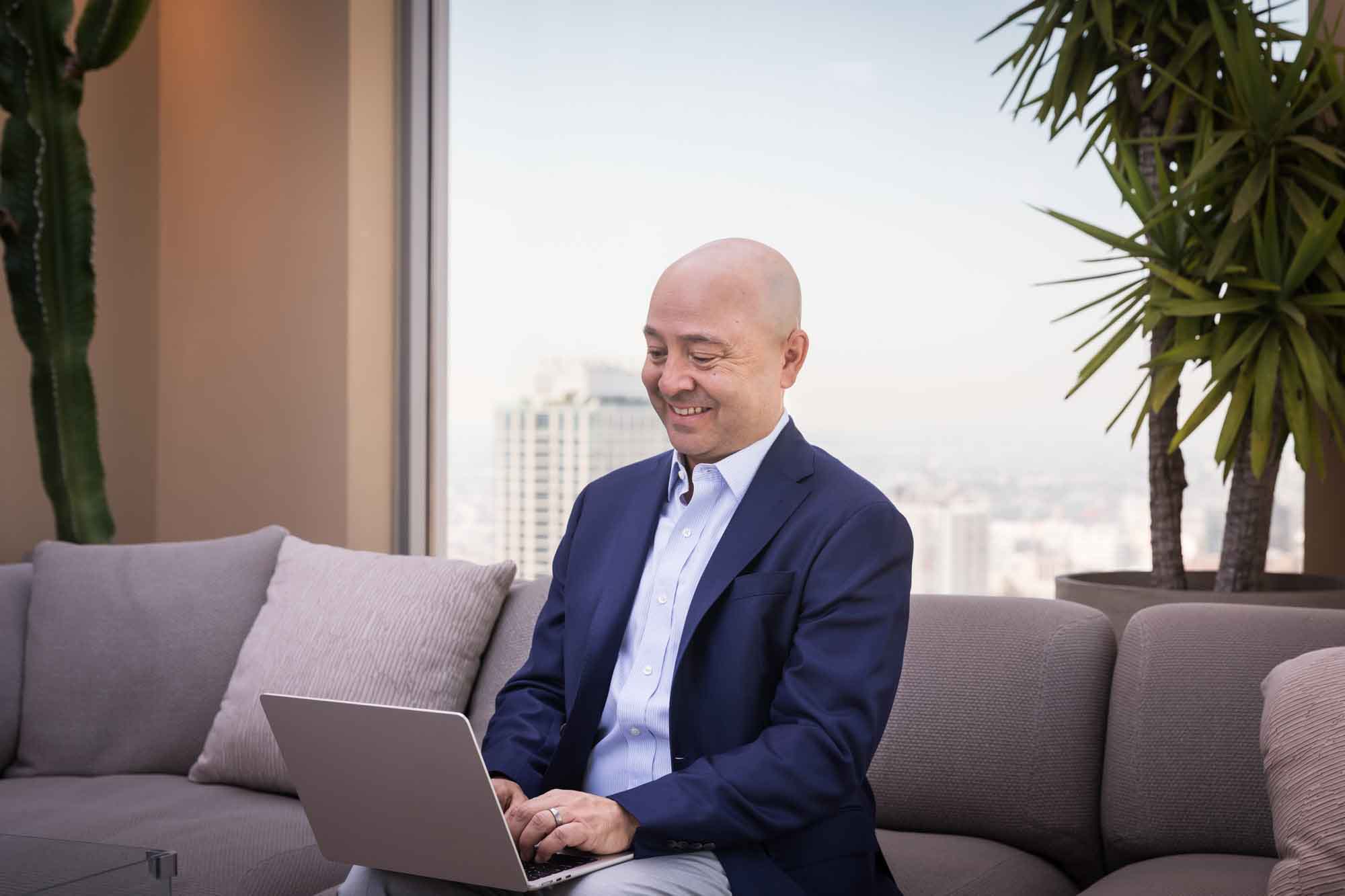 Executive portrait of businessman wearing blue blazer and blue and white shirt seated on grey couch typing on laptop in front of window for an article on how to prepare for a branding photo shoot