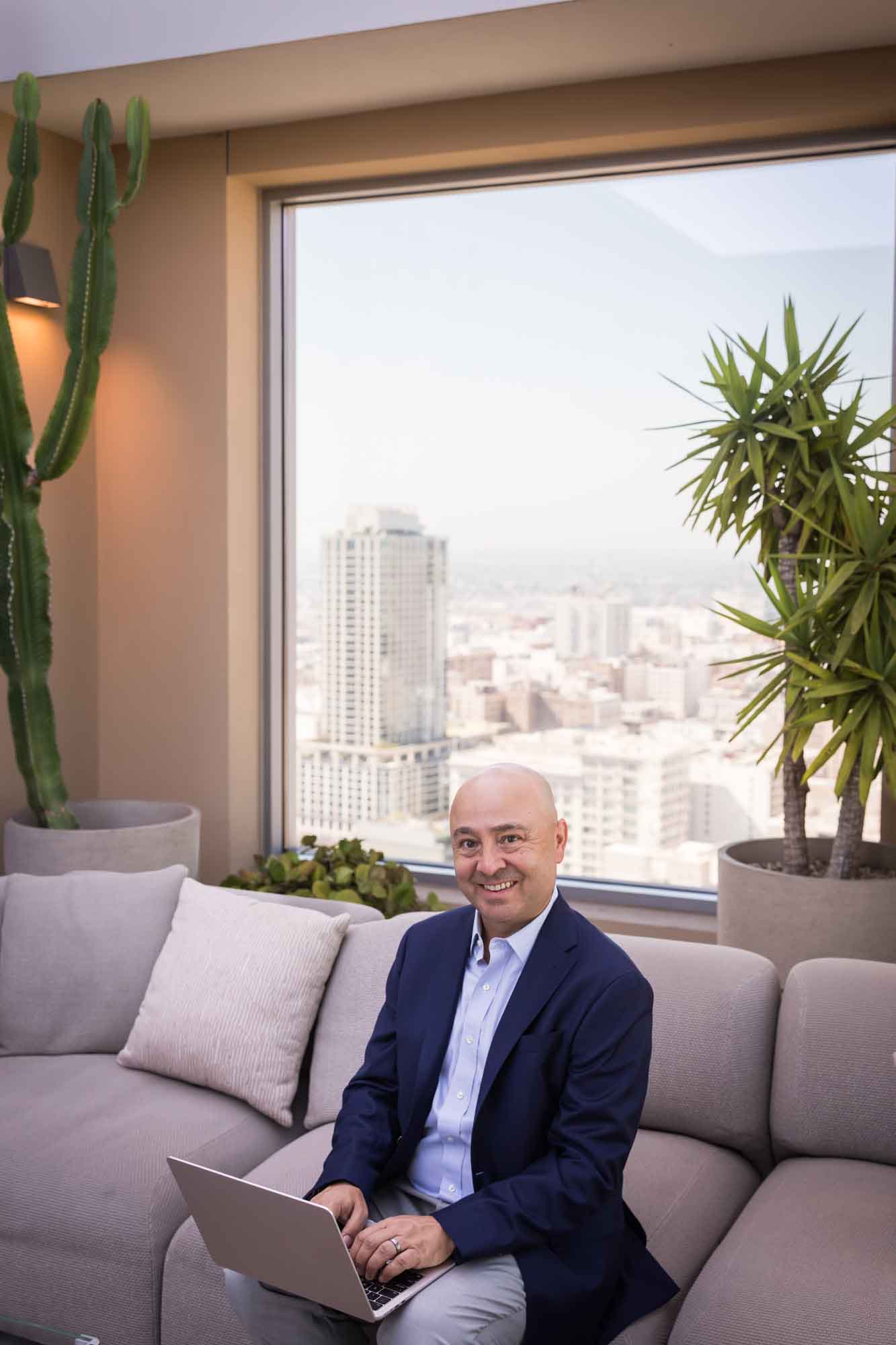 Executive portrait of businessman wearing blue blazer and blue and white shirt seated on grey couch in front of window for an article on how to prepare for a branding photo shoot
