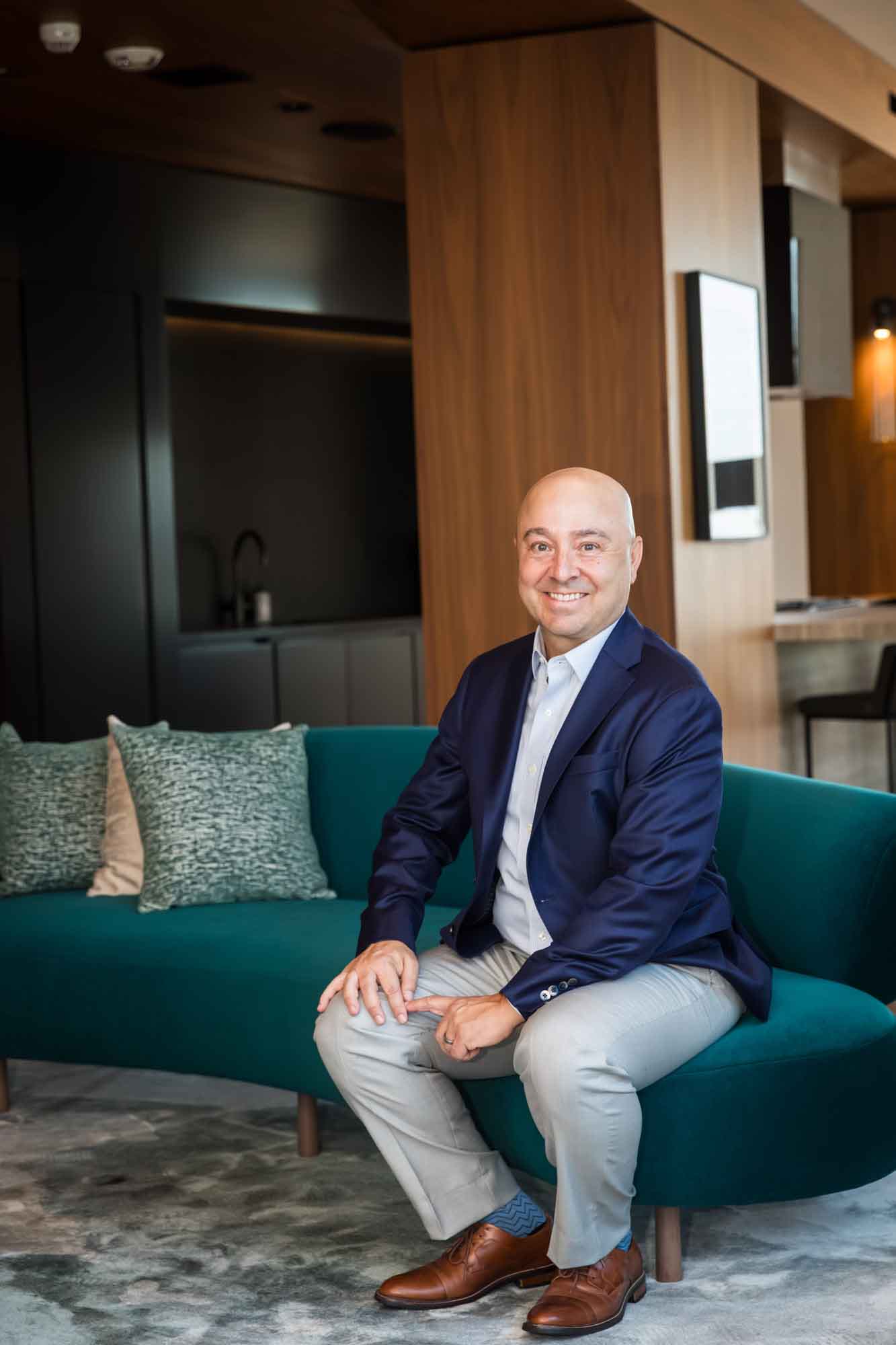 Executive portrait of businessman wearing blue blazer and blue and white shirt seated on teal couch for an article on how to prepare for a branding photo shoot