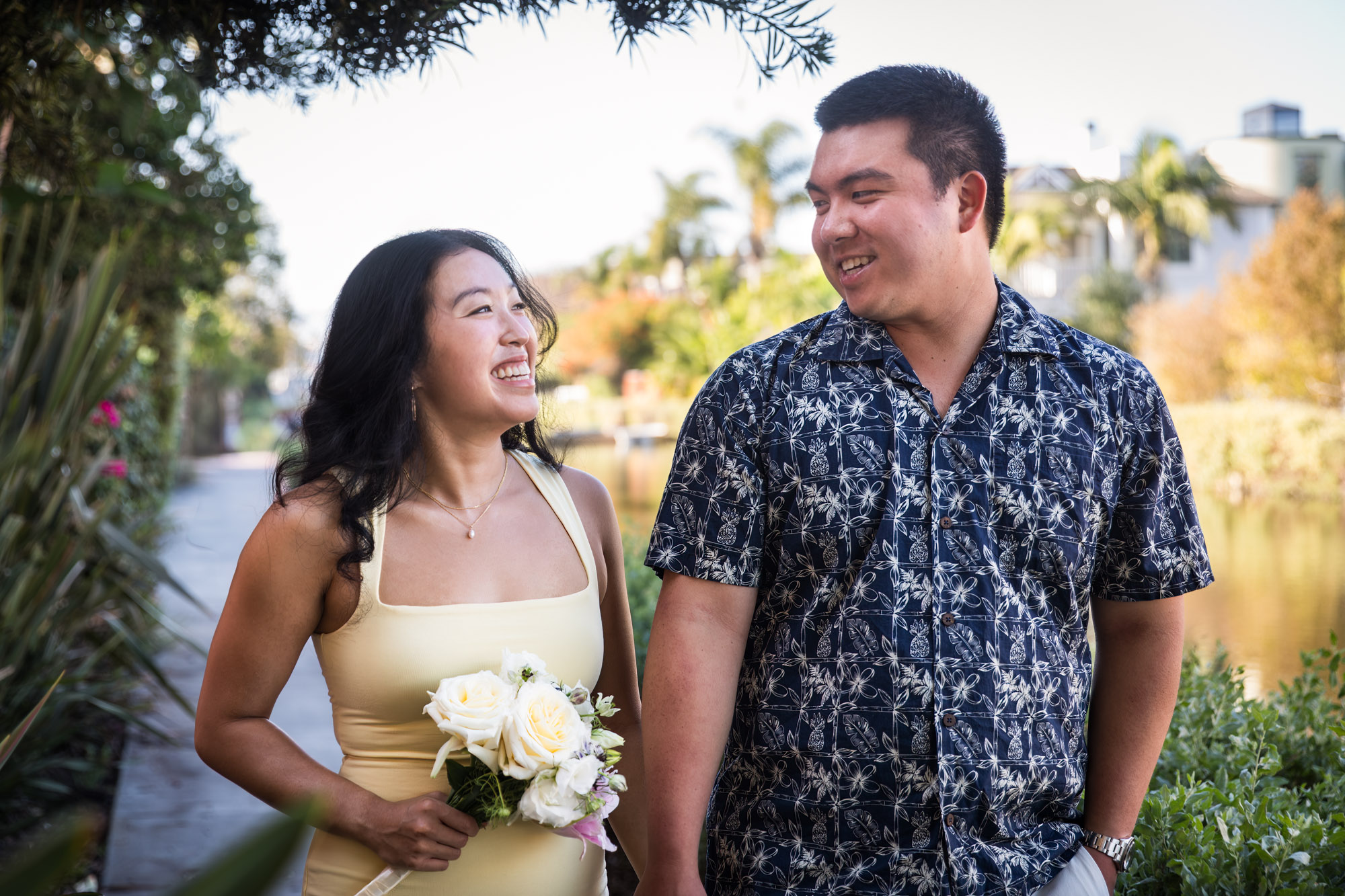 Woman wearing long yellow dress walking hand-in-hand with man wearing blue and white patterned shirt along pathway beside canal with house in background during a Venice Canals engagement photo shoot