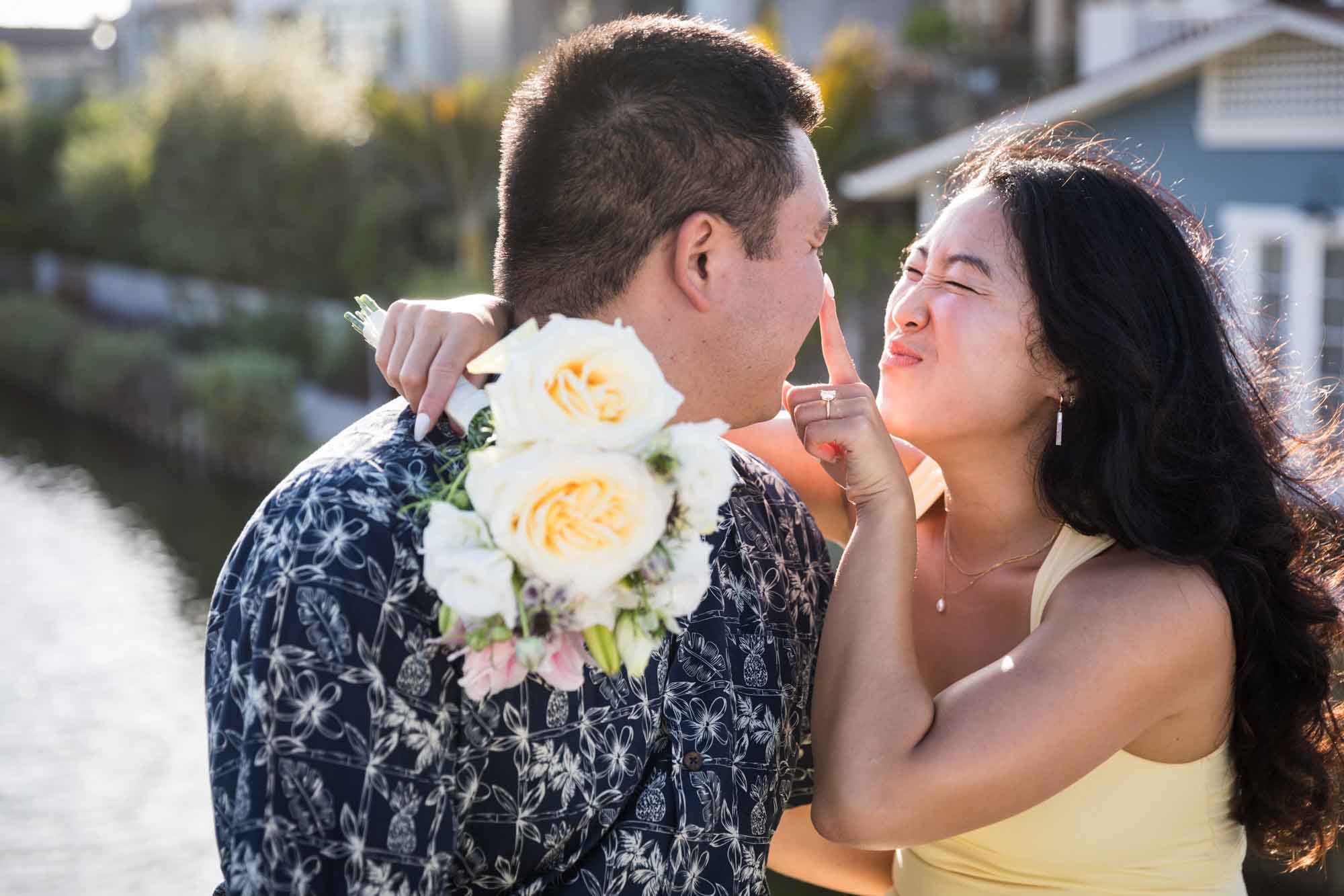 Woman wearing sleeveless yellow dress touching nose of man wearing blue and white patterned shirt with canals in background during a Venice Canals engagement photo shoot