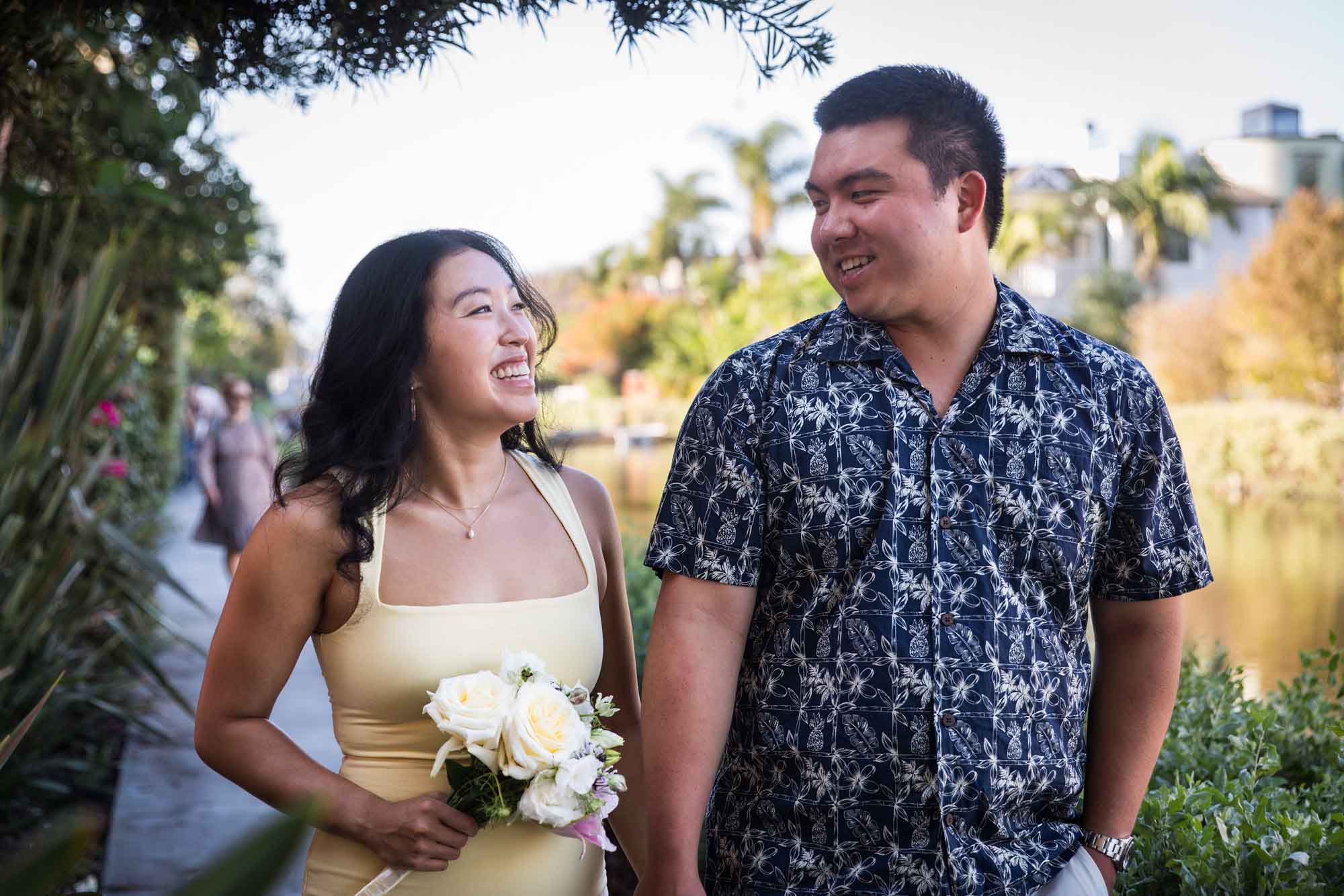 Woman wearing long yellow dress walking hand-in-hand with man wearing blue and white patterned shirt along pathway beside canal with house in background during a Venice Canals engagement photo shoot