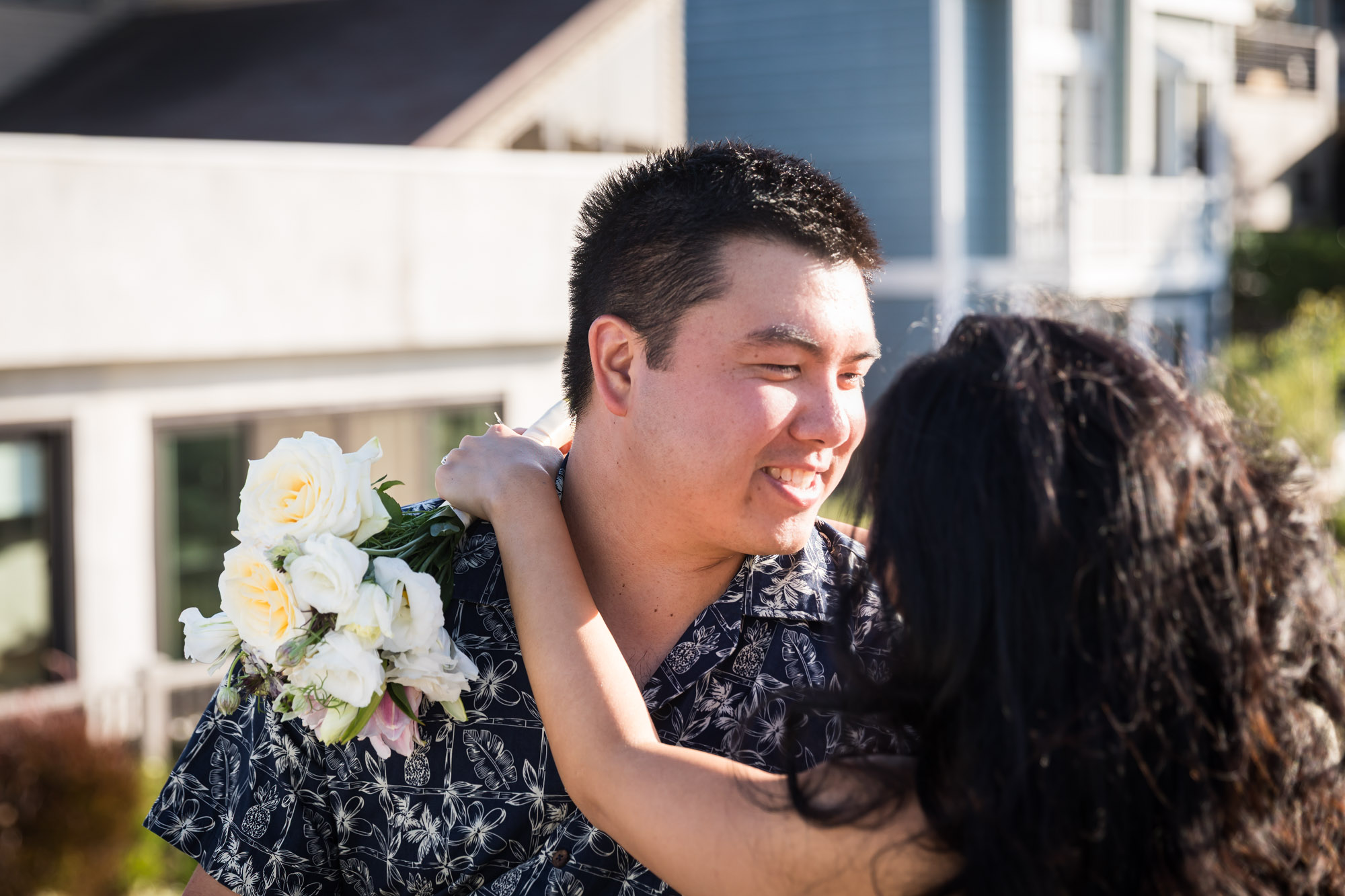 Woman wearing sleeveless yellow dress sitting in lap of man wearing blue and white patterned shirt with canals in background during a Venice Canals engagement photo shoot