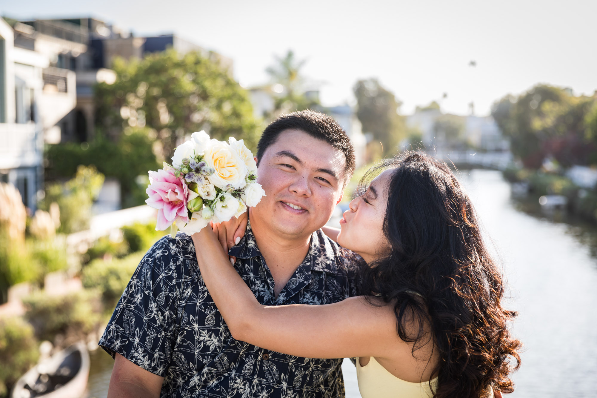 Woman wearing sleeveless yellow dress sitting in lap of man wearing blue and white patterned shirt with canals in background during a Venice Canals engagement photo shoot