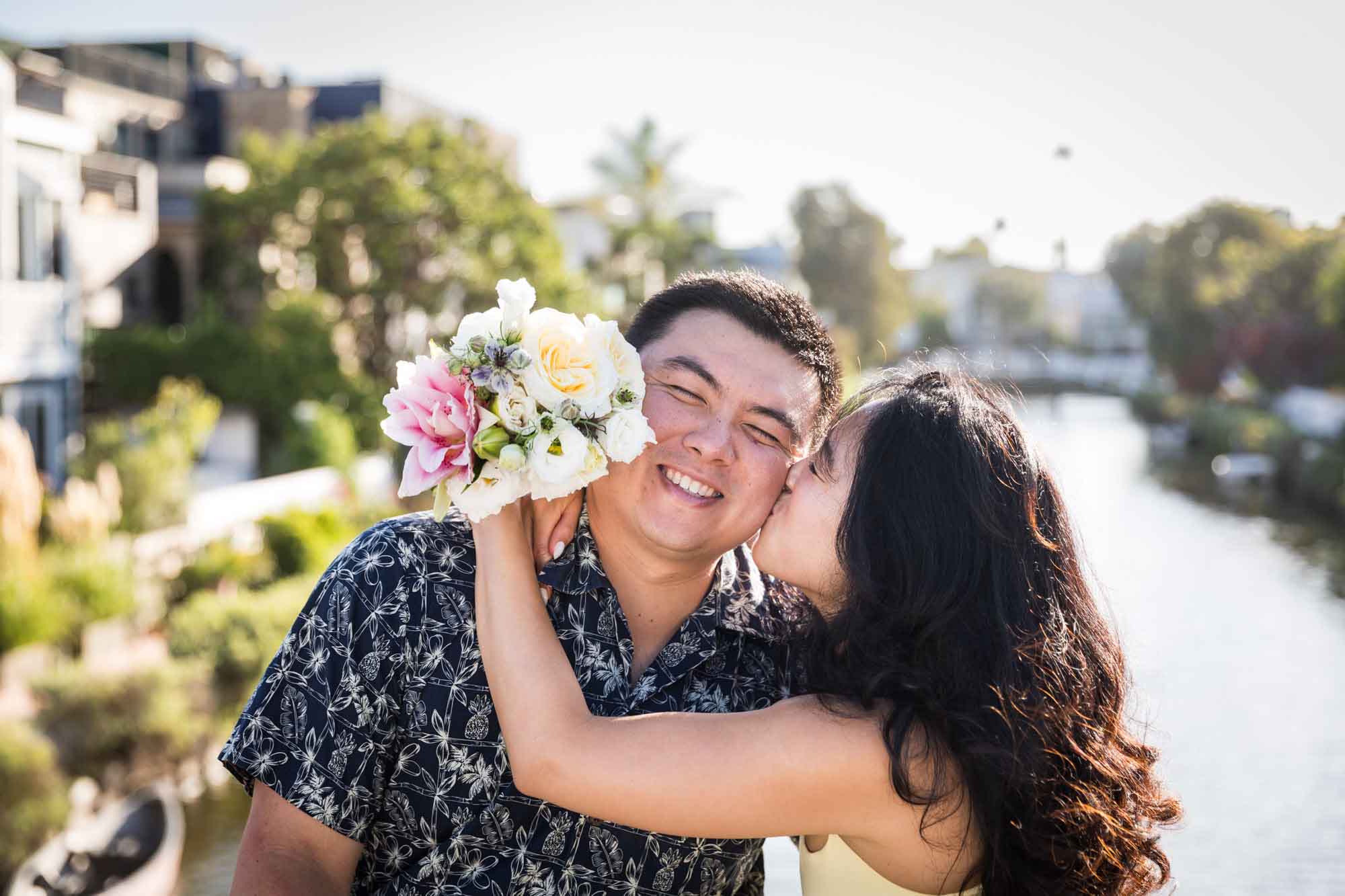 Woman wearing sleeveless yellow dress kissing man wearing blue and white patterned shirt with canals in background during a Venice Canals engagement photo shoot