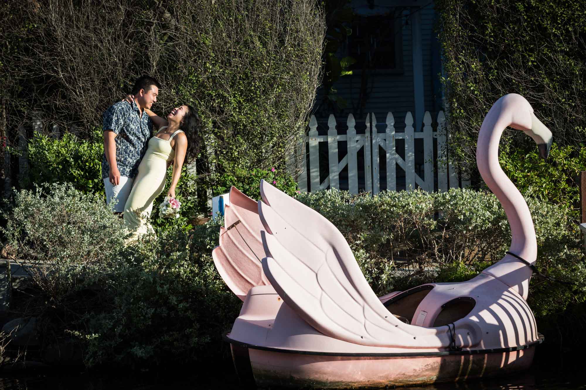 Woman wearing sleeveless yellow dress holding flowers standing next to man wearing blue and white patterned shirt with canals and pink flamingo float in background during a Venice Canals engagement photo shoot