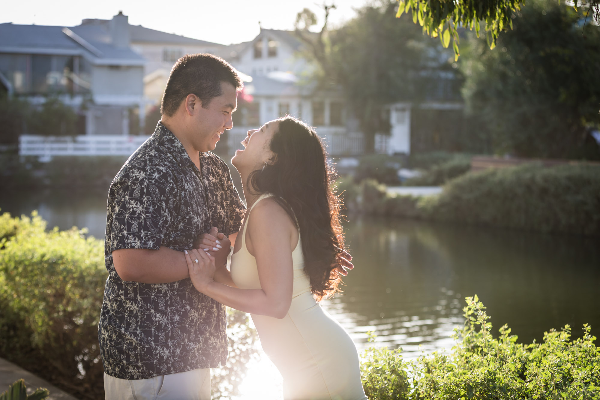 Woman wearing sleeveless yellow dress holding hands with man wearing blue and white patterned shirt with canals in background during a Venice Canals engagement photo shoot