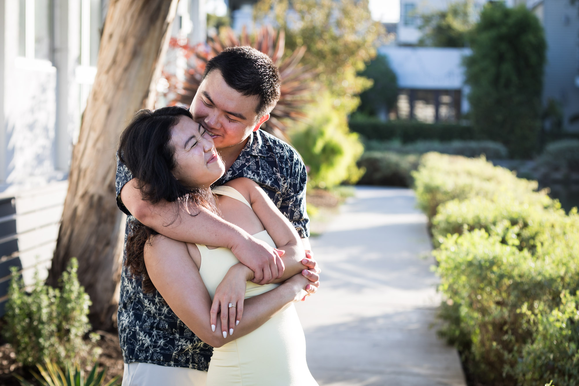Man wearing blue and white patterned shirt hugging woman wearing sleeveless yellow dress from behind with with canals in background during a Venice Canals engagement photo shoot