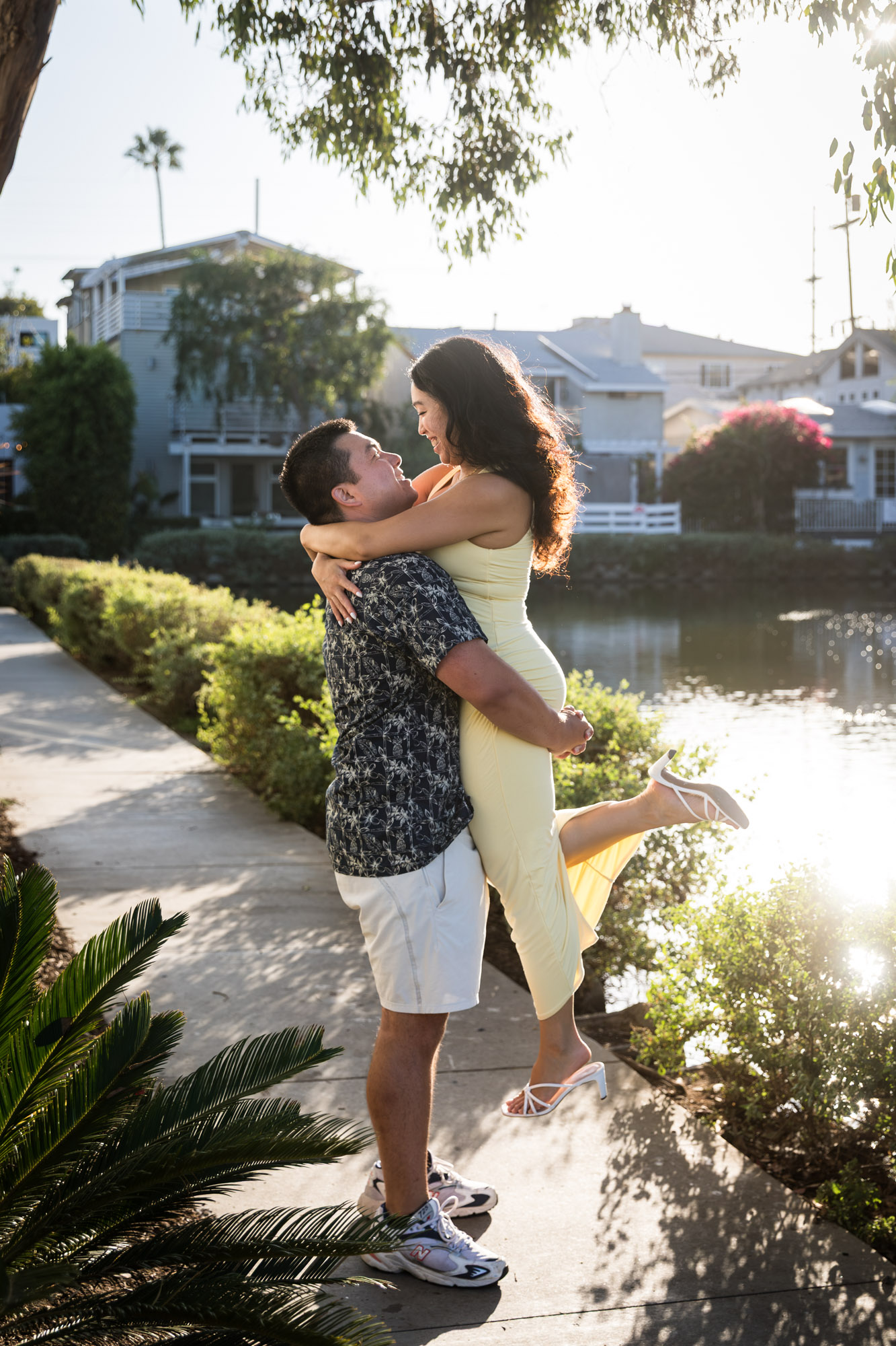 Man wearing blue and white patterned shirt lifting woman wearing sleeveless yellow dress in the air with canals in background during a Venice Canals engagement photo shoot