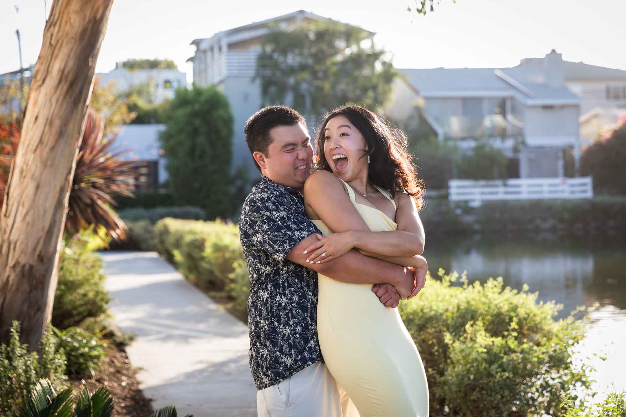 Man wearing blue and white patterned shirt hugging woman wearing sleeveless yellow dress from behind with with canals in background during a Venice Canals engagement photo shoot