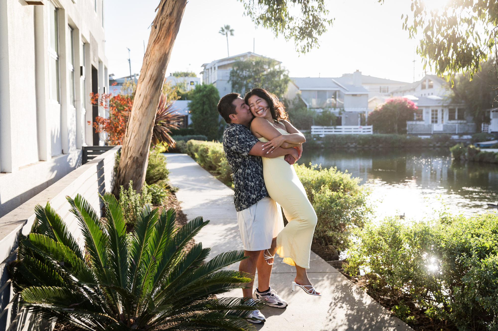 Man wearing blue and white patterned shirt hugging woman wearing sleeveless yellow dress from behind with canals in background during a Venice Canals engagement photo shoot