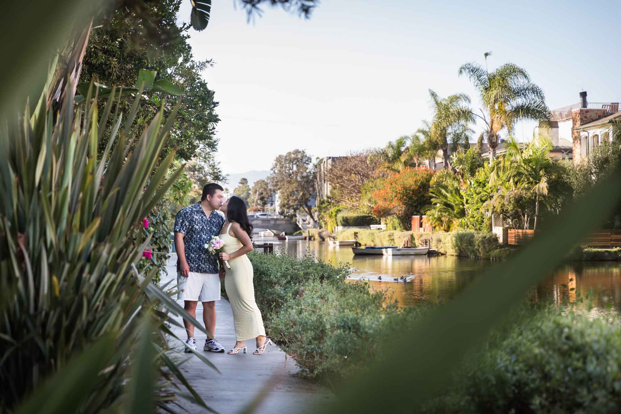 Woman wearing long yellow dress walking hand-in-hand with man wearing blue and white patterned shirt along pathway beside canal with house in background during a Venice Canals engagement photo shoot