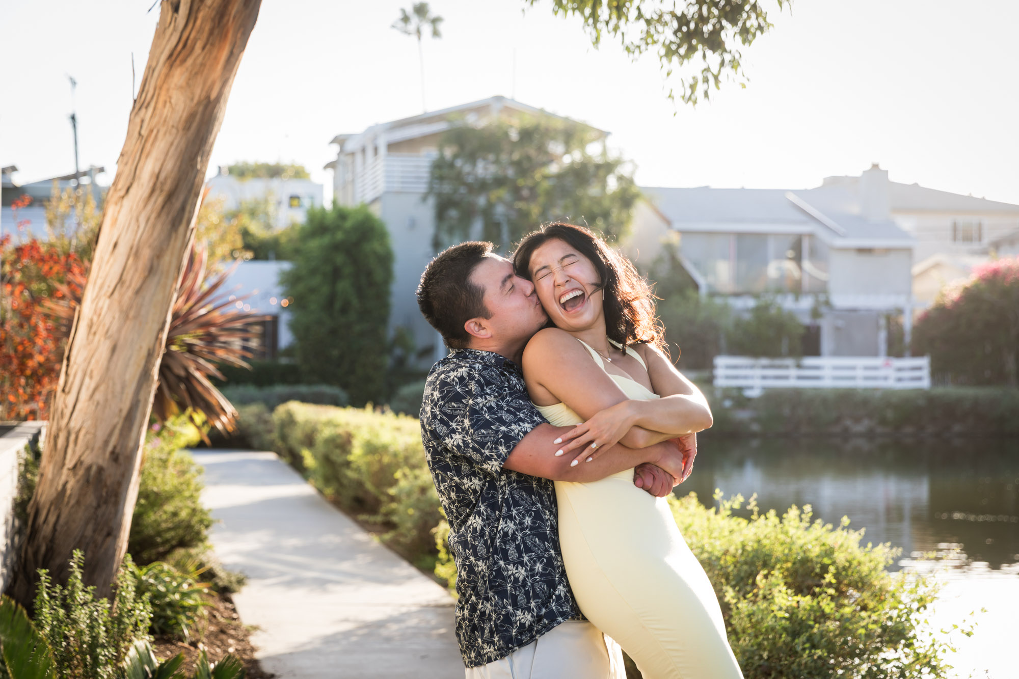 Man wearing blue and white patterned shirt hugging woman wearing sleeveless yellow dress from behind with canals in background during a Venice Canals engagement photo shoot